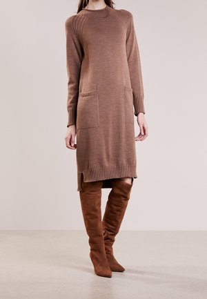 Žena oblečená v hnědých úpletových šatech s dlouhým rukávem a předními kapsami, doplněných hnědými semišovými kozačkami ke kolenům, stojící před jednobarevným pozadím.