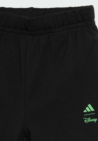 Čierne športové nohavice z jemného materiálu s elastickým opaskom. Obsahujú zelené logo adidas a Disney. Hladká textúra.