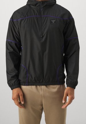 Man draagt een zwarte hoodie met paarse biezen en beige broek, armen ontspannen langs zijn zij, kijkt recht vooruit.