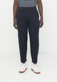 Σκούρο μπλε φόρμα με παχιά ελαστική μέση, σχεδιασμό με στενή γραμμή στα πόδια και λείο, απαλό ύφασμα. Φοριέται με λευκά αθλητικά παπούτσια.