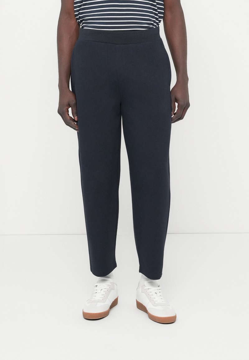 Σκούρο μπλε φόρμα με παχιά ελαστική μέση, σχεδιασμό με στενή γραμμή στα πόδια και λείο, απαλό ύφασμα. Φοριέται με λευκά αθλητικά παπούτσια.