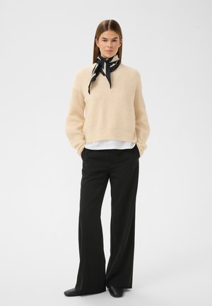 Krēmkrāsas adīts džemperis ar gariem piedurknēm, kopā ar melnām, plaša piegriezuma biksēm. Apkārt kaklam sasaistīts melns un balts šalle, kas piešķir detaļu.