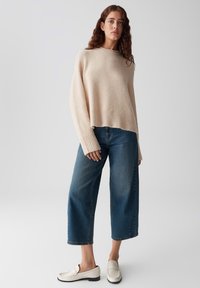 Donna con capelli ricci che indossa un maglione oversized beige, jeans a gamba larga blu e mocassini crema, in piedi contro uno sfondo grigio semplice.