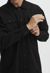 Zwarte geknoopte blouse van katoen, met een puntige kraag, twee borstzakken en lange mouwen met knoopmanchetten.