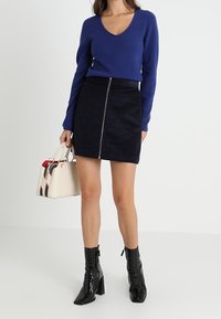 Pull en maille bleu à col en V, associé à une mini-jupe en suède noire avec une fermeture éclair à l'avant. Accessoires comprenant un sac à main crème et des bottes à talons noires.