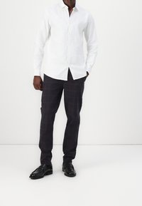 Chemise blanche à boutons faite d'un tissu léger, associée à un pantalon à carreaux sombres avec un motif en quadrillage rouge et des chaussures noires.