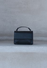 Sac à main noir en cuir embossé façon croco avec poignée supérieure posé sur une marche en béton contre un mur gris uni.
