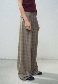 Pantalon à jambes larges à carreaux dans des tons de brun avec un tissu texturé. Il présente des plis à la taille et des chaussures noires à enfiler avec des accents.