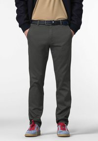 Man in een donkergrijze broek, met een zwarte gevlochten riem, een beige shirt, een marineblauwe jas en blauwe sneakers met rode veters, staande met handen in de zakken.
