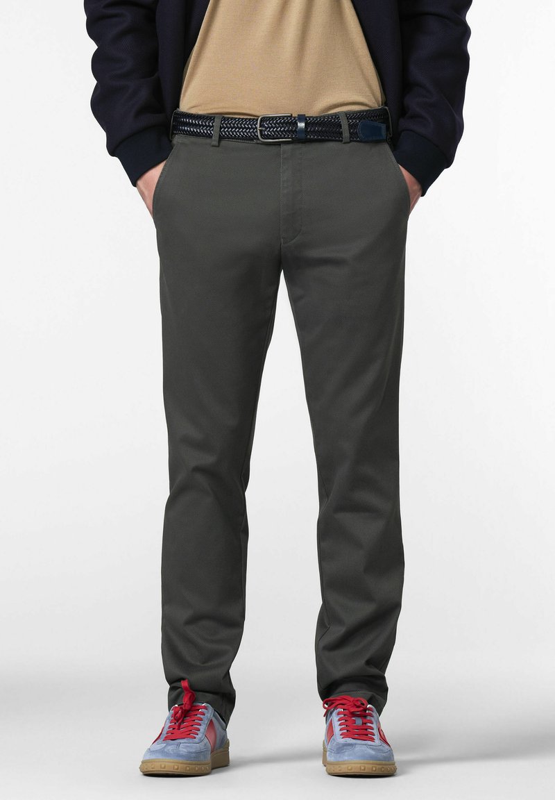 Man in een donkergrijze broek, met een zwarte gevlochten riem, een beige shirt, een marineblauwe jas en blauwe sneakers met rode veters, staande met handen in de zakken.