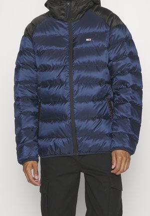 Giacca a vento blu navy con design trapuntato, finitura lucida e dettagli neri sulle spalle; presenta una chiusura frontale con zip e cappuccio regolabile.