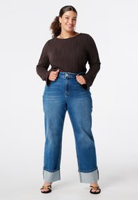 Haut long à manches brunes texturé avec un motif en vagues, associé à un jean en denim bleu clair taille haute avec des poignets retroussés et une coupe décontractée.