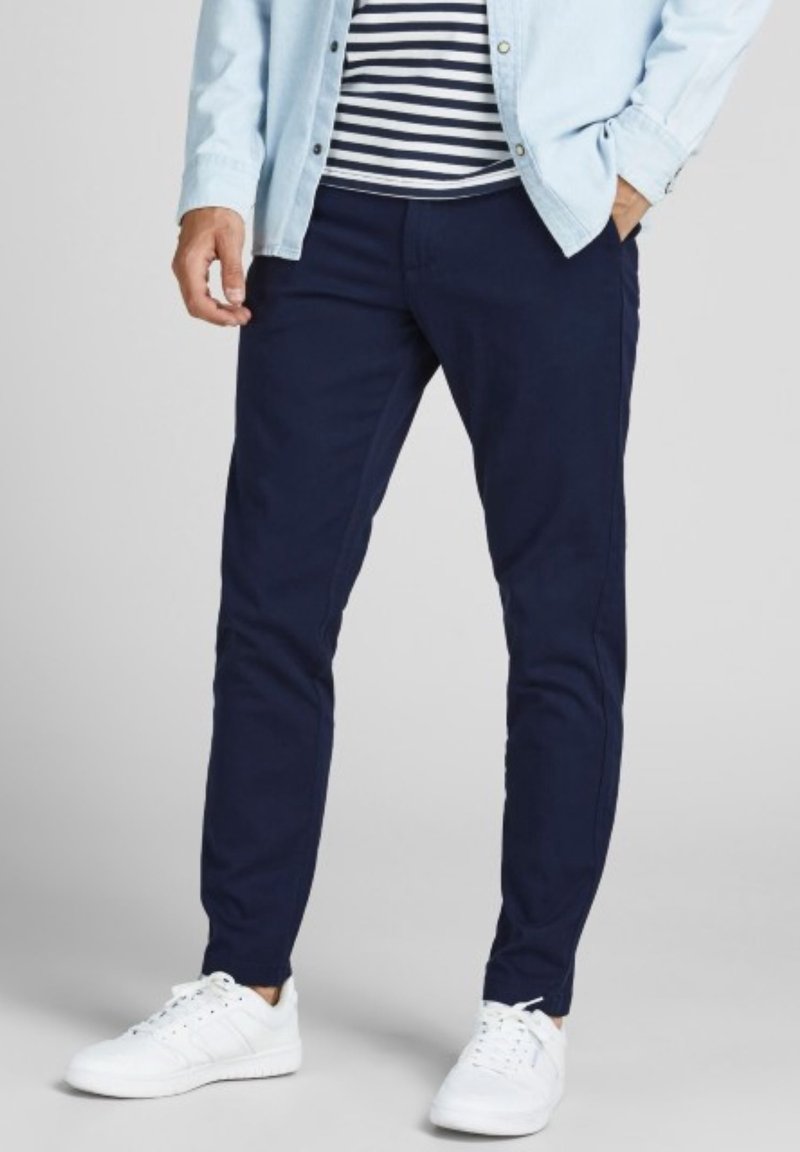 Homme portant un pantalon bleu marine, des baskets blanches, une chemise rayée et une chemise bleu clair à boutons, debout avec une main dans la poche.