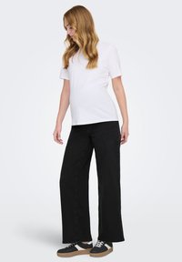 Gravid kvinde med langt, bølget hår iført en hvid kortærmet top, sorte bukser med vide ben og sorte og hvide sneakers, der kigger ned.