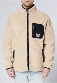 Beige Fleecejacke mit hohem Kragen, kontrastierender schwarzer Tasche und Reißverschluss vorne. Verfügt über strukturierten Stoff und ein kleines Markenlabel.