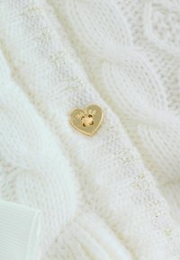 Weißer Strickpullover mit strukturiertem Muster, ausgestattet mit einem goldenen herzförmigen Charm, auf dem "Baker" eingraviert ist, und einem subtilen Schimmer.
