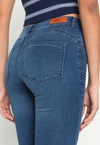 Jeans de ganga azul com um design ajustado, apresentando uma etiqueta de couro marcada com "ONLY" e dois bolsos traseiros com detalhes de costura visíveis.
