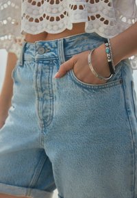 Lichtblauwe denimshorts met een hoge taille, voorzien van een knoopsluiting, opgerolde hem en zijvakjes. Vergezeld van zilveren en turquoise armbanden.