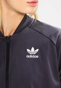Svart idrottsjacka i slät tyg, med dragkedja och vit adidas-logotyp på vänster bröst. Detaljerad sömnad på axlarna.