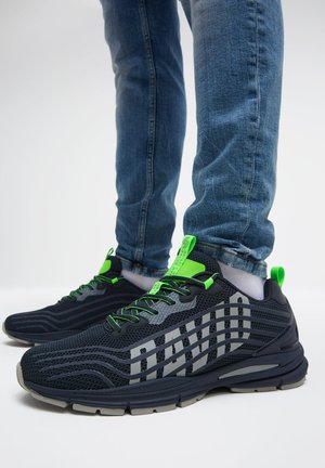 Person, die schwarze und graue Sportschuhe mit neongrünen Akzenten, blaue Jeans und weiße Socken vor weißem Hintergrund trägt.