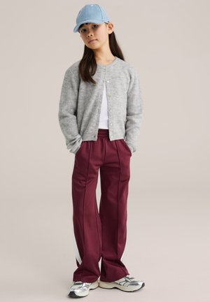 Mladá osoba nosiaca svetlomodrú šiltovku, sivý kardigán, biele tričko, bordové tepláky s bielymi pruhmi a strieborné tenisky, stojaca s rukami v vreckách.