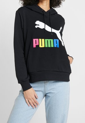 Person trägt schwarzen Hoodie mit weißem springendem Puma-Logo und buntem "PUMA"-Text, kombiniert mit hellblauen Jeans.