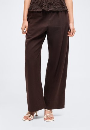 Kvinde iført mørkebrune vidde bukser, en tekstureret mørk top og hvide remsandaletter med hæl, stående mod en ensfarvet lys baggrund.