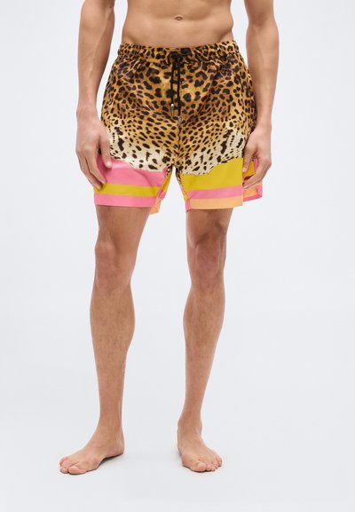Homme portant un short à cordon avec un haut imprimé léopard et des blocs de couleur rose et jaune, debout pieds nus devant un fond uni.