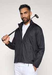 Melnas vieglas virsjakas ar rāvējslēdzēja priekšpusi, stāvus apkakli un sānu kabatām, uzvilkta pār baltu polo krekls.