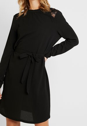 Femme portant une robe noire à manches longues avec des détails en dentelle sur les épaules et une ceinture nouée à la taille, debout avec une main sur la hanche.
