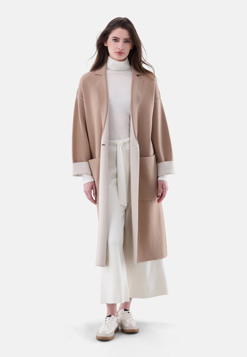 Beige long coat with a collar, oversized shape, and light cream cuffs. Worn over a white turtleneck and wide-leg white pants with sneakers.