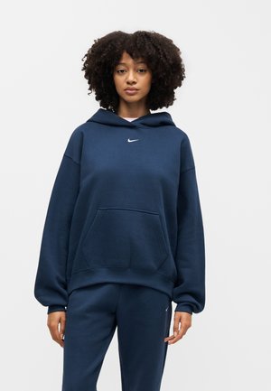 Mladi žena kovrčave kose, odjevena u tamnoplavi Nike duks s kapuljačom i odgovarajuće trenirke, stoji ispred jednostavne svijetle pozadine.