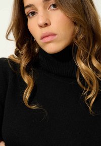 Czarny sweter z kołnierzem typu turtle neck o prążkowanej strukturze, z wysokim dekoltem i dopasowanym krojem. Częściowo widoczne kręcone brązowe włosy.