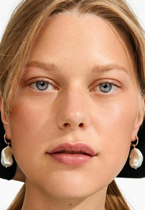 Pärlilaternaga kõrvarõngad kuldse rõngaga, millel on ebaregulaarsed kujundid ja läikiv valge viimistlus, kantud naturaalse meigi ja lahtiste juustega.