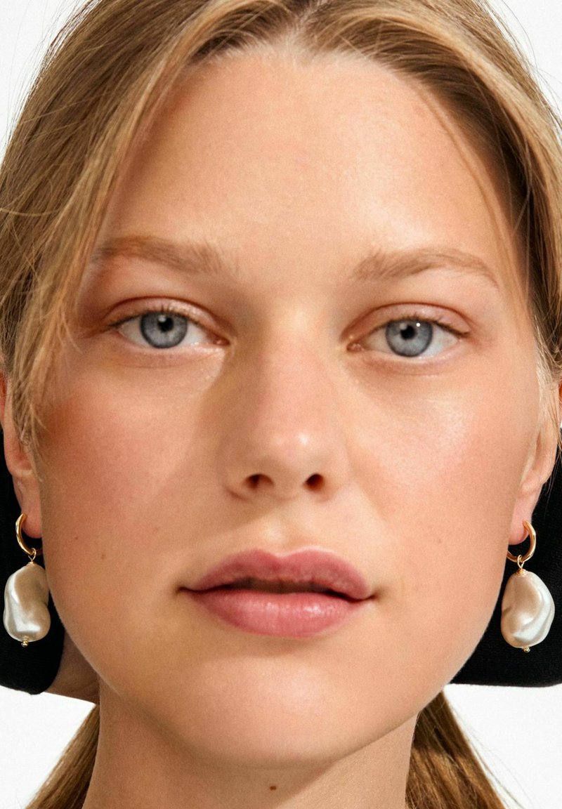 Kolczyki z perłami i złotą obręczą, o nieregularnych kształtach i błyszczącym białym wykończeniu, noszone z naturalnym makijażem i rozpuszczonymi włosami.