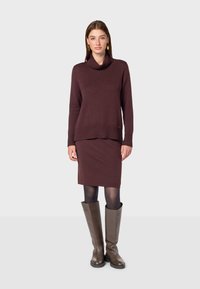 Weinrote Strickjacke mit einem Kollerkragen, gerippten Bündchen und Saum, kombiniert mit einem passenden weinroten Rock. Getragen mit kniehohen braunen Lederstiefeln.