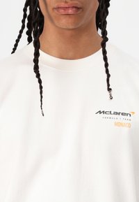 Hvid bomulds t-shirt med sorte og orange logoer: "McLaren," "FORMULA 1 TEAM," og "MONACO." Rund hals og korte ærmer.