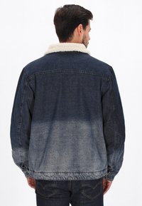 Sininen farkkutakki, jossa on vaalea sherpa-kaulus, tummansininen liukuväri, nappikiinnitys ja rento istuvuus. Tekstuuri näyttää pehmeältä ja kuluneelta.