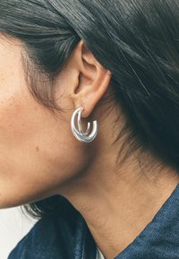 Orecchini a cerchio in argento con un design ondulato, caratterizzati da due curve intrecciate. La finitura è lucida e gli orecchini sono di dimensioni medie.