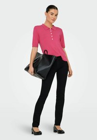 Donna in piedi che indossa una polo rosa a maniche corte, pantaloni neri slim, scarpe nere con tacco basso, mentre porta una grande borsa nera a tracolla.