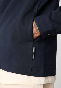 Hand in navy blue long-sleeve shirt pocket, showing tattoo on wrist and fabric tag reading "Marc O'Polo" near hem.