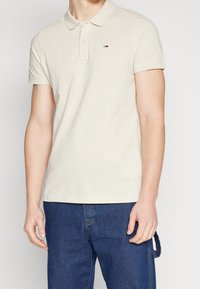 Beige Polo-Shirt mit kurzen Ärmeln und kleinem Flaggenlogo auf der Brust, kombiniert mit dunkelblauen Jeans und einer seitlichen Denim-Schlaufe.