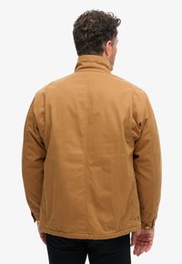 Chaqueta de algodón marrón con cuello alto, textura acolchada y costuras traseras. Presenta acentos de botones en los puños y un ajuste relajado.