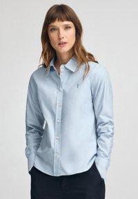 Kvinna med ljusbrunt hår klädd i en ljusblå skjorta med knappar och en liten broderad logotyp, stående med händerna i fickorna mot en enfärgad bakgrund.