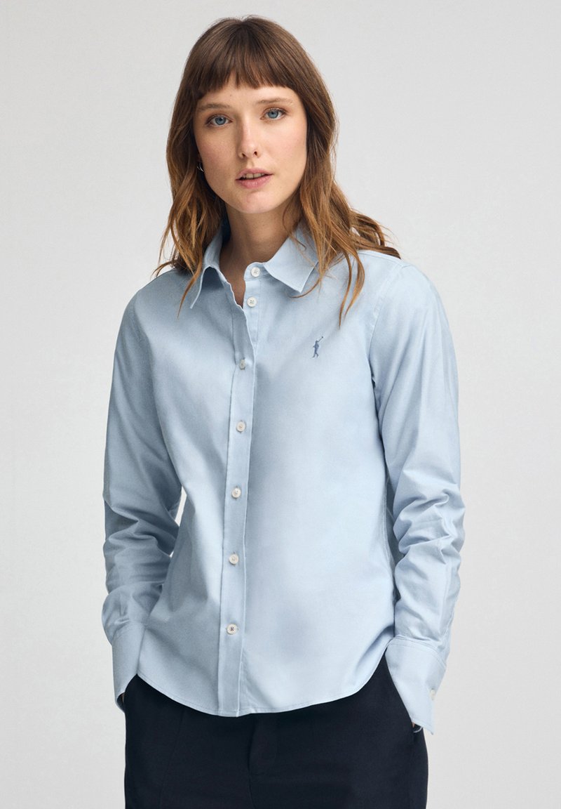 Kvinna med ljusbrunt hår klädd i en ljusblå skjorta med knappar och en liten broderad logotyp, stående med händerna i fickorna mot en enfärgad bakgrund.