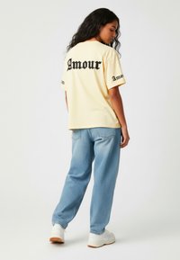 Lichtgele oversized t-shirt met zwarte "Amour" tekst op de achterkant en mouwen, gecombineerd met lichtblauwe denim jeans en witte sneakers.