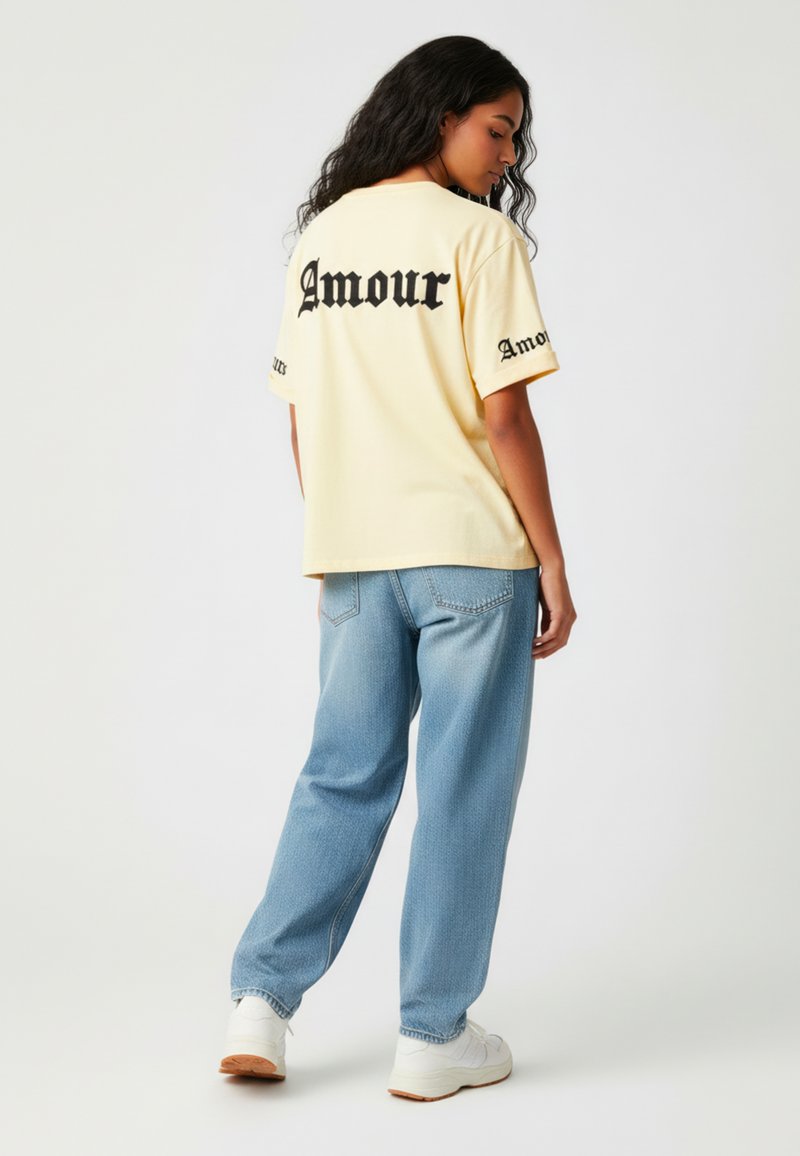 Lichtgele oversized t-shirt met zwarte "Amour" tekst op de achterkant en mouwen, gecombineerd met lichtblauwe denim jeans en witte sneakers.