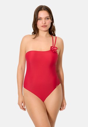 Mujer con un traje de baño rojo de un solo hombro con un detalle floral en la tira, posando frente a un fondo claro y liso.