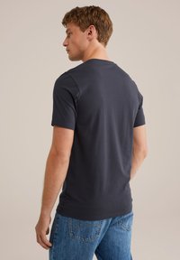Tânăr stând deoparte, purtând un tricou simplu gri închis și blugi albaștri, pe fundal bej.