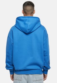 Kék kapucnis pulóver laza szabással, bordázott mandzsettákkal és szegéllyel. Sima textúrájú, kenguruzsebbel és túlméretezett kapucnival.